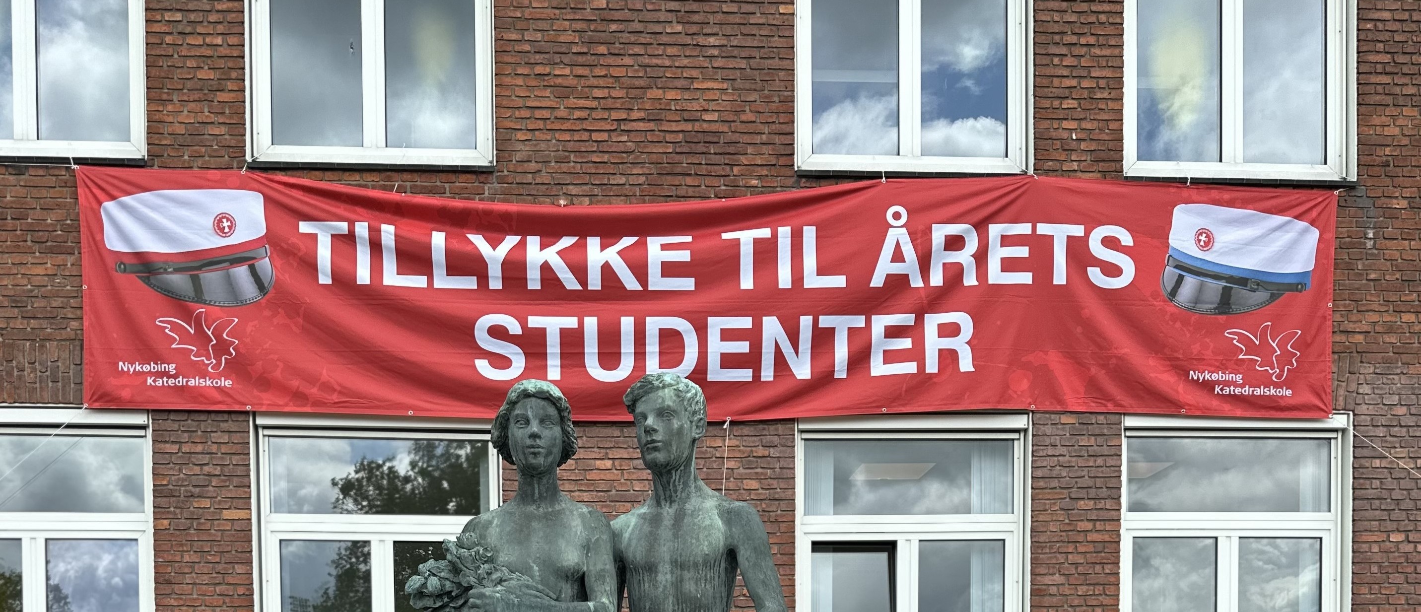 Rødt banner på skolens mur med teksten Tillykke til årets studenter samt stx-hue i venste side og hf-hue i højre side