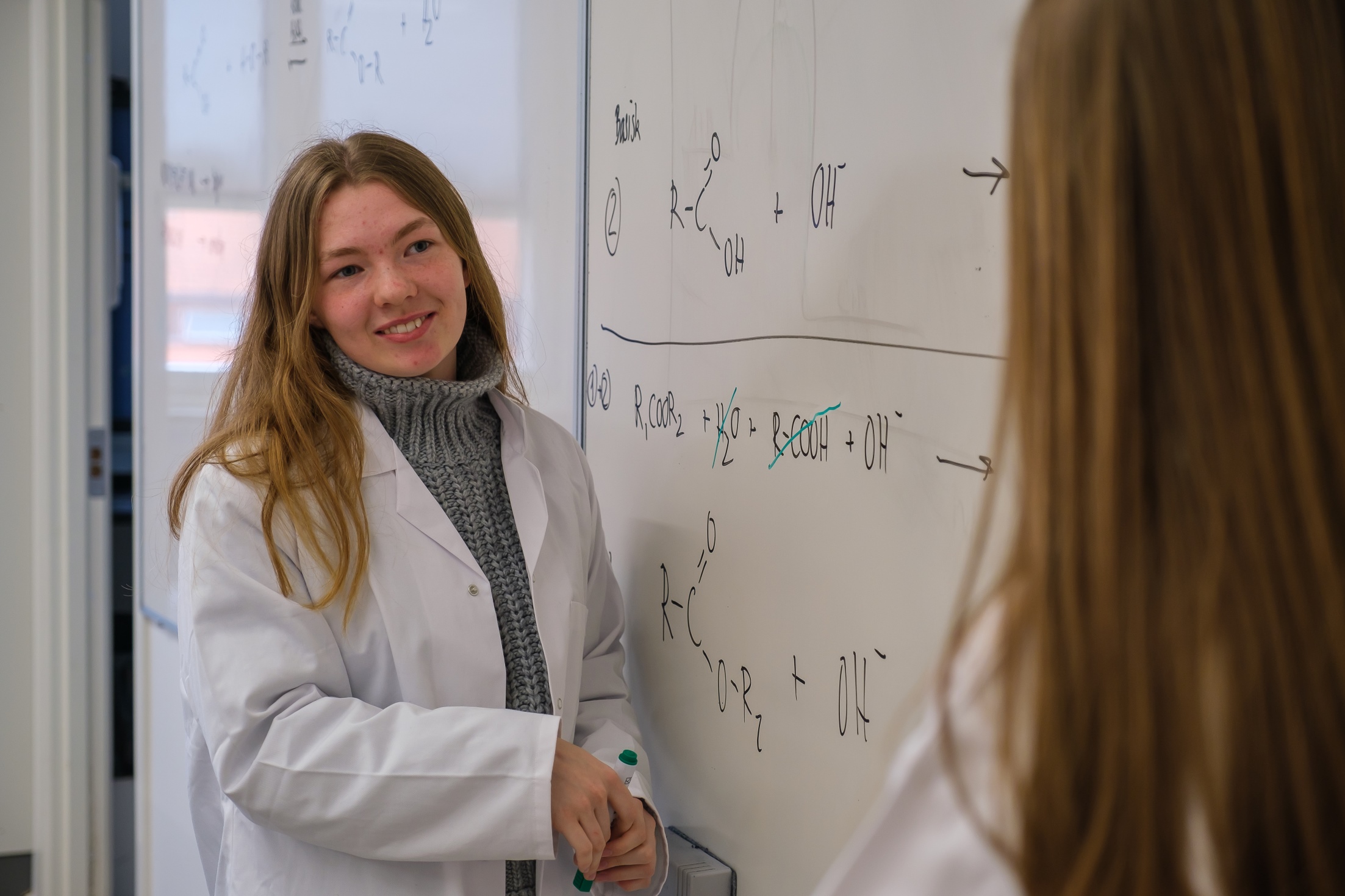 Pige i hvid kittel skriver formler på tavlen i kemilokale