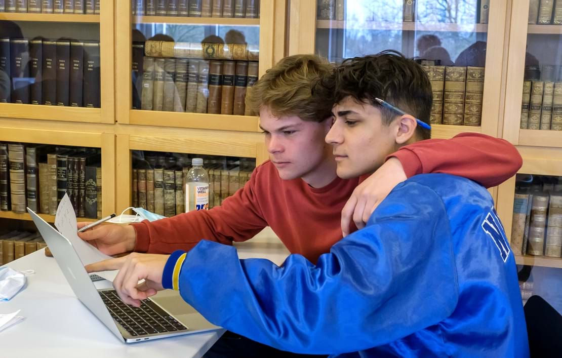 To drenge sidder og kigger på computerskærm foran skabe med bøger